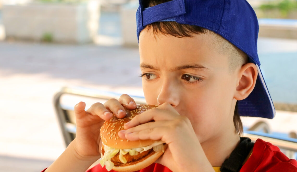 Ein Kind mit blauer Baseballkappe isst draußen einen Burger